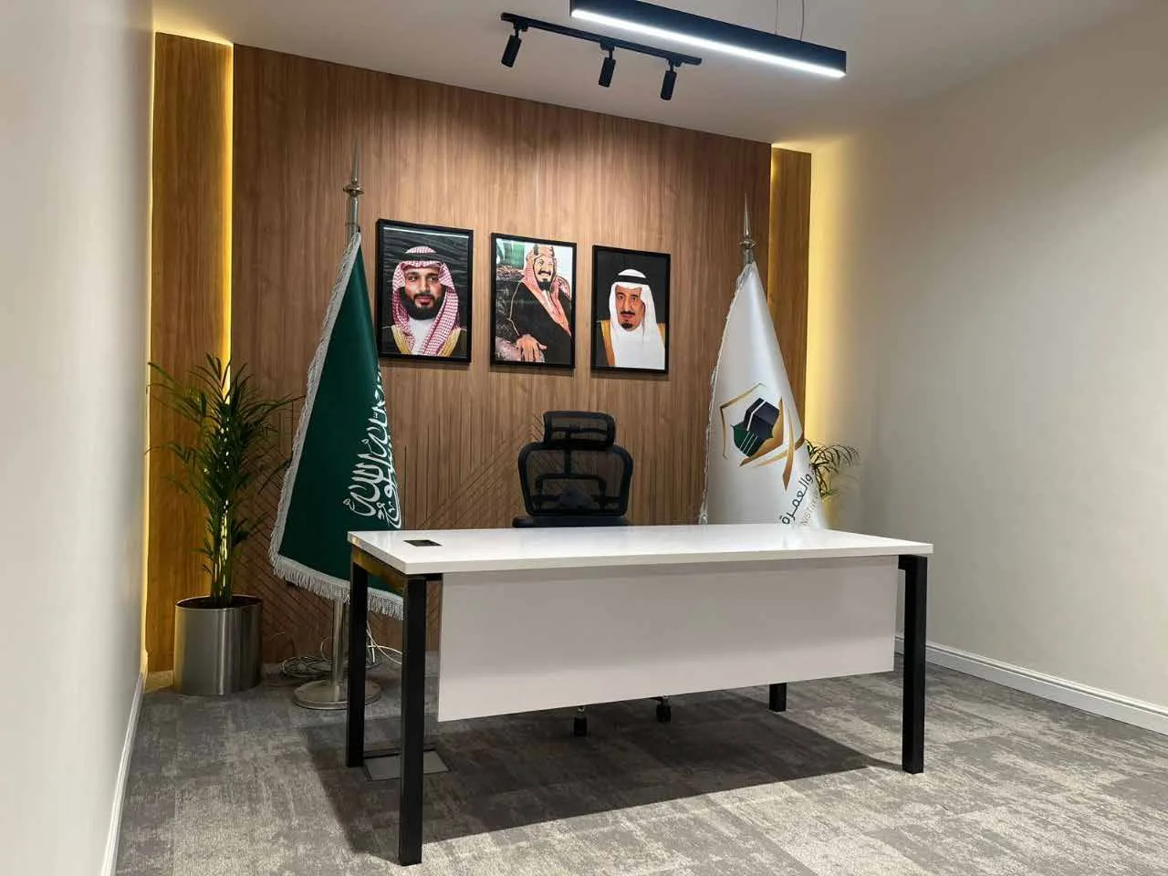 مكتب تنفيذي بطاولة بيضاء وكرسي عمل أسود وجدار مكسو بالخشب وإطارات معلقة